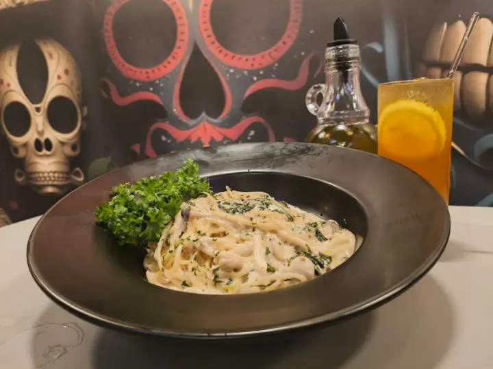 Creamy pasta plate garnished with parsley beside an amber drink, vibrant skull mural behind